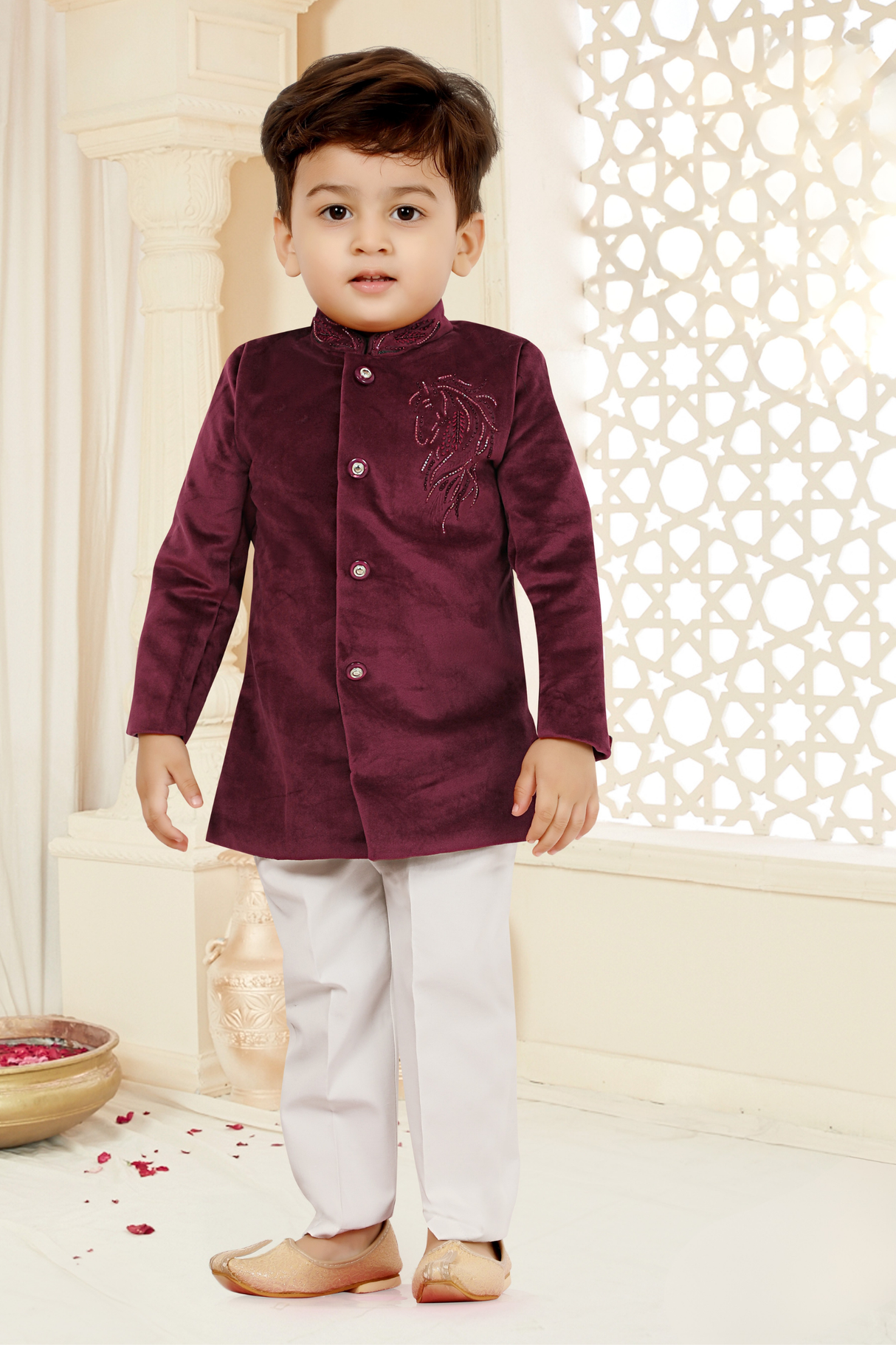 Toddler boy wearing a deep maroon/burgundy velvet short tailored jacket with silver horse embroidery on the chest, paired with cream trousers and gold ethnic shoes.