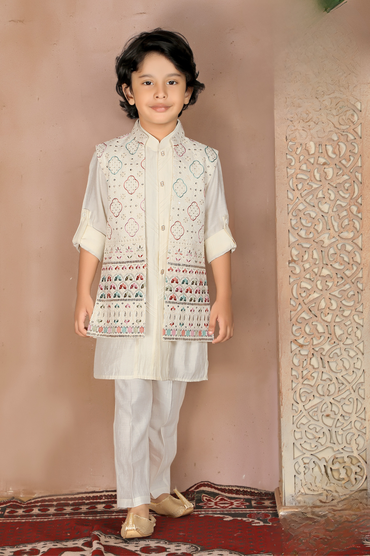 Young boy wearing a fawn-colored kurta pajama set, layered with a sleeveless Nehru jacket featuring a repeated pattern of multi-colored elephants, and light ethnic slip-on shoes.