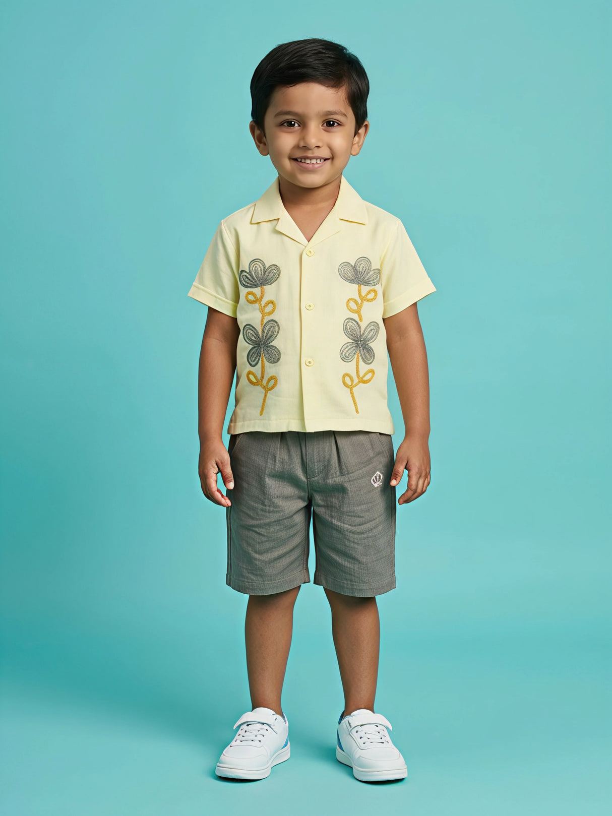 A young boy wearing a pastel yellow short-sleeve Cuban collar shirt with grey and yellow floral embroidery on the front, paired with matching grey casual shorts and white sneakers, set against a solid turquoise background as seen in image_3bf8a7.jpg.