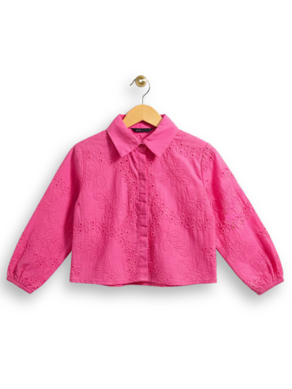 Front view of a girls' bright hot pink long-sleeve button-up blouse completely covered in delicate white eyelet (broderie anglaise) embroidery, hanging on a wooden hanger.