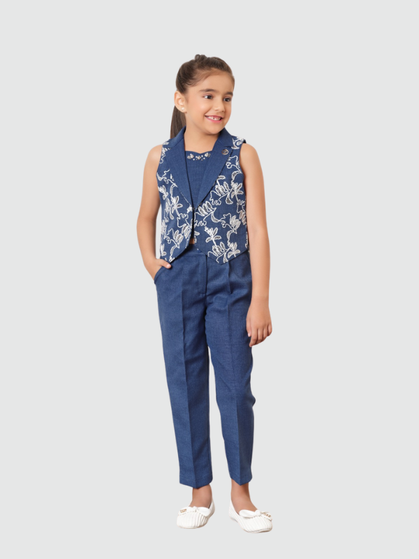 Young girl smiling in a sleeveless blue denim-look waistcoat with white floral embroidery, paired with matching tailored blue trousers and white ballet flats