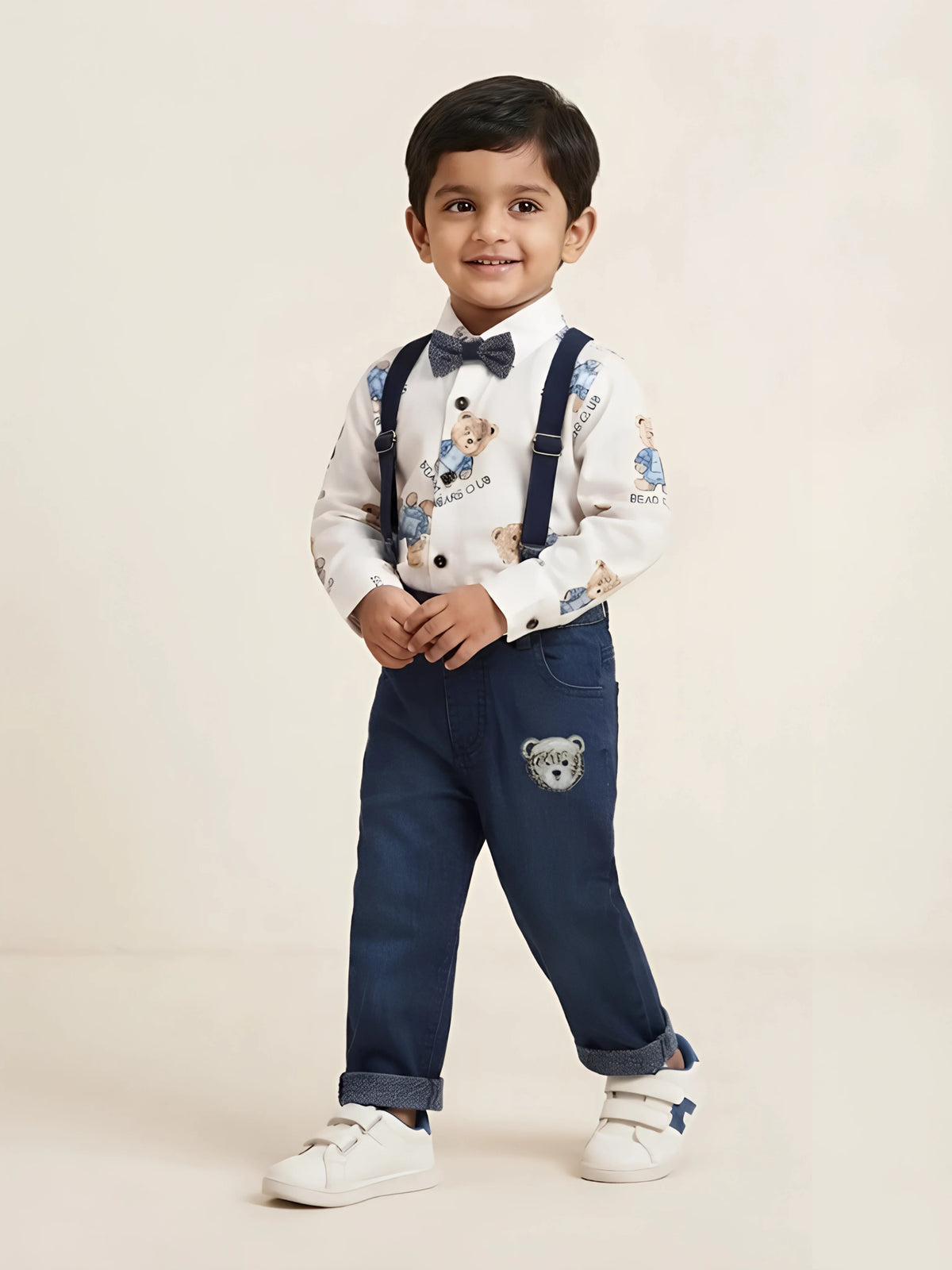 Young boy wearing a white full-sleeve shirt with teddy bear prints, a bow tie, navy suspenders, and dark blue denim trousers