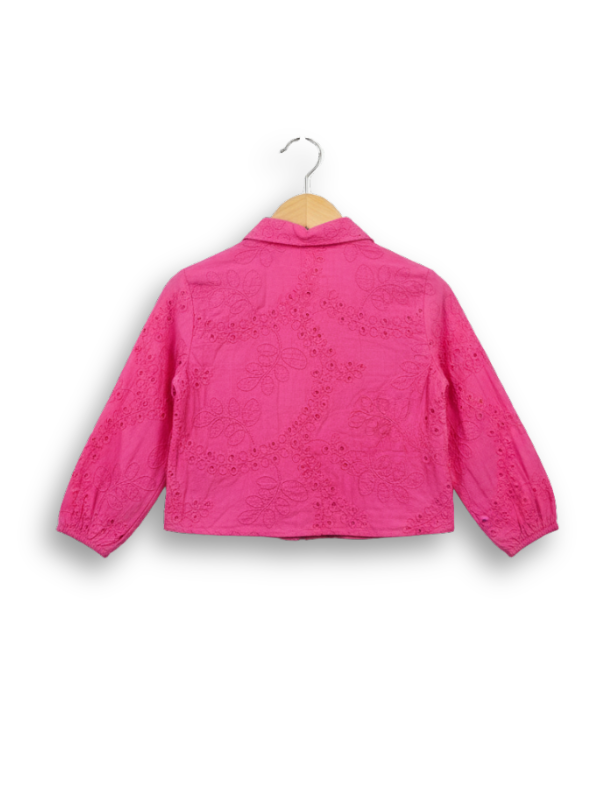 Front view of a girls' bright hot pink long-sleeve button-up blouse completely covered in delicate white eyelet (broderie anglaise) embroidery, hanging on a wooden hanger.
