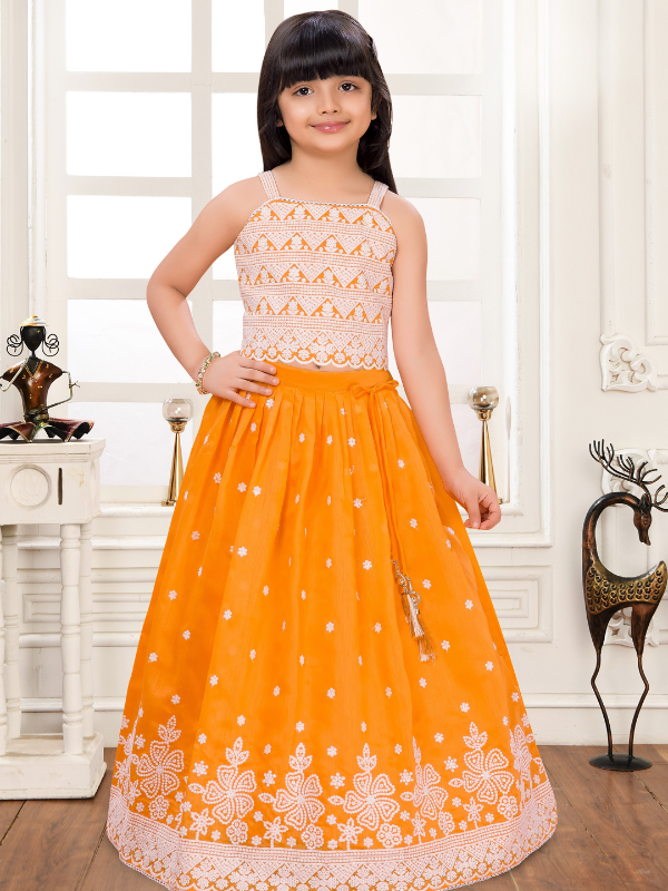 Young girl wearing a sleeveless orange and white lehenga choli set. The top is white with orange patterned embroidery, and the flared orange skirt features a thick white floral embroidered border.