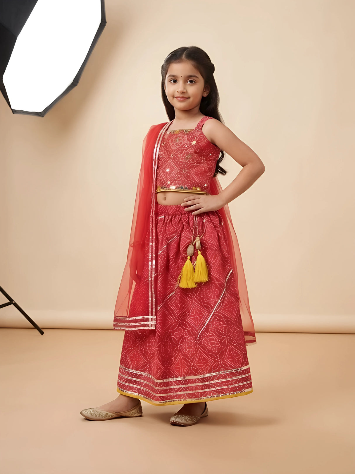 Young girl posing in a vibrant red printed lehenga choli set with gold gota patti border, yellow tassels, and sheer red dupatta