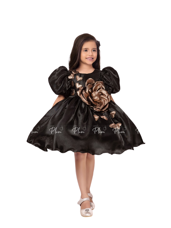 Young girl smiling in a black satin party dress with large puff sleeves and a metallic gold 3D rose accent on the bodice.