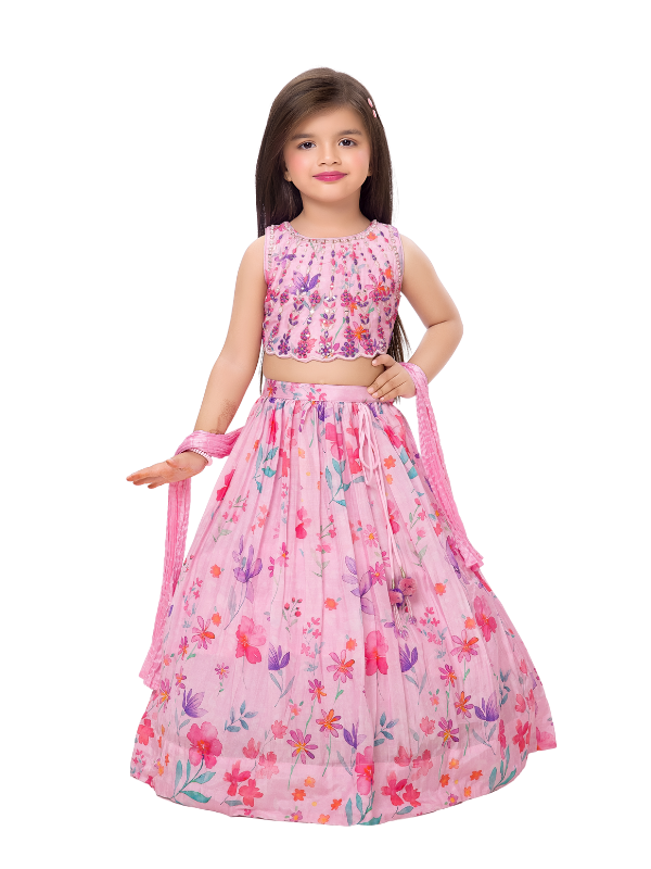 A young girl modeling a beautiful pink lehenga choli set with a sequin-embroidered crop top and a flared skirt featuring a bright, multi-color floral watercolor print, holding a matching pink dupatta.