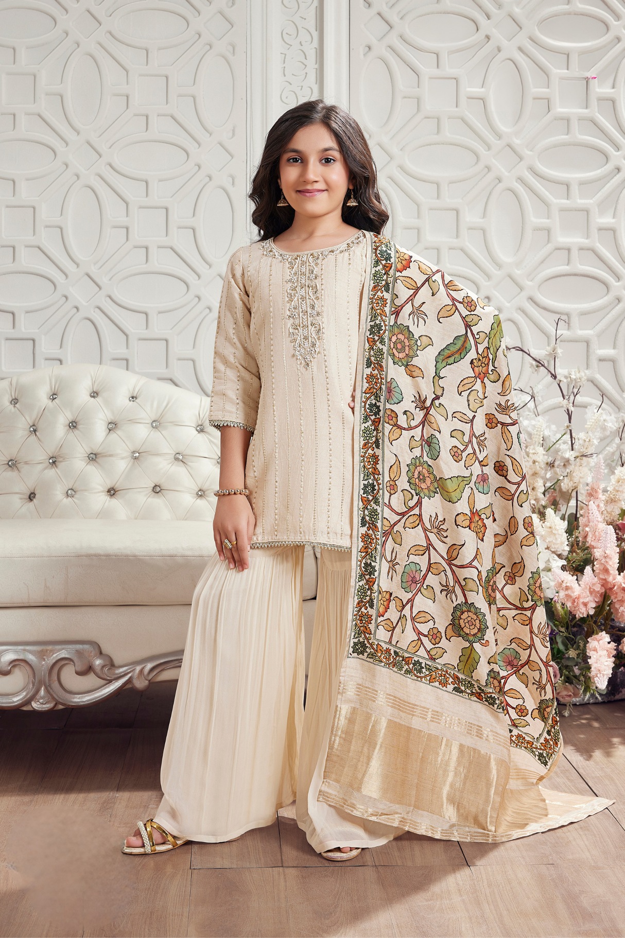 Young girl wearing an elegant cream gharara suit, with a sequin-embroidered short kurti, a pleated gharara, and a large dupatta with a colorful floral border.