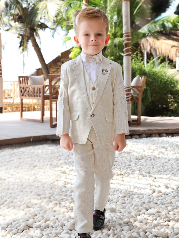 Full-body shot of a young boy wearing a light cream linen-blend suit (jacket, vest, pants) and a matching bow tie, standing outdoors on a sunny, pebbled path in a resort-like setting.
