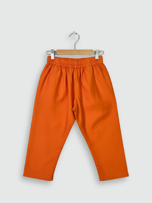 A young boy standing and smiling, wearing a bright orange long-sleeved kurta and pajama set, accessorized with a sleeveless teal, orange, and cream geometric-printed Nehru jacket.