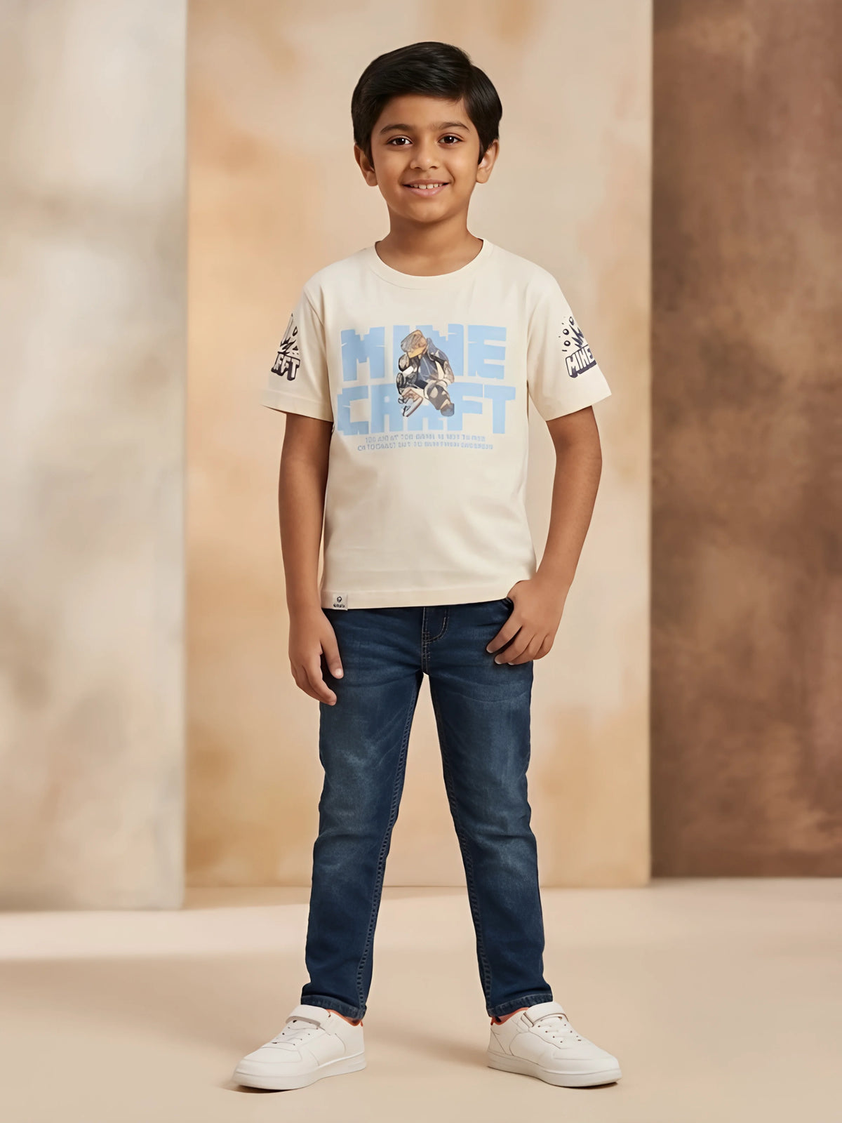 Young boy wearing a cream short-sleeve t-shirt with blue graphic print on front and sleeves, paired with denim jeans