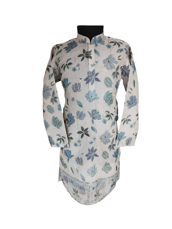 Full-length image of a young boy wearing a cream/off-white traditional kurta with all-over blue floral embroidery and a matching border, paired with white churidar pajamas and gold ethnic shoes.