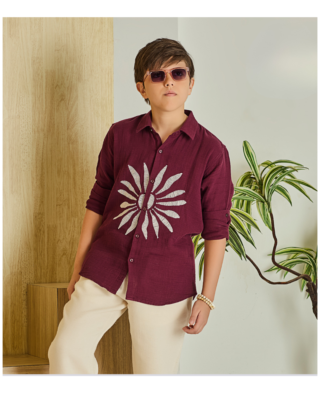 Young boy wearing a maroon/burgundy button-down shirt with a large white flower appliqué on the chest, sleeves rolled up, and light cream trousers.
