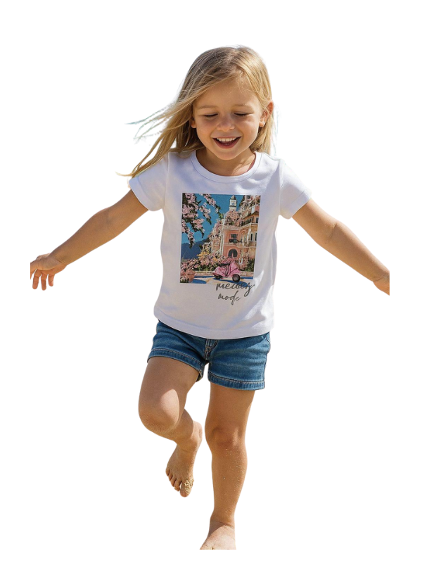 Smiling young girl running outdoors wearing a short-sleeve white t-shirt with a colorful graphic print of a European street scene and the words "holiday mode", paired with denim shorts.