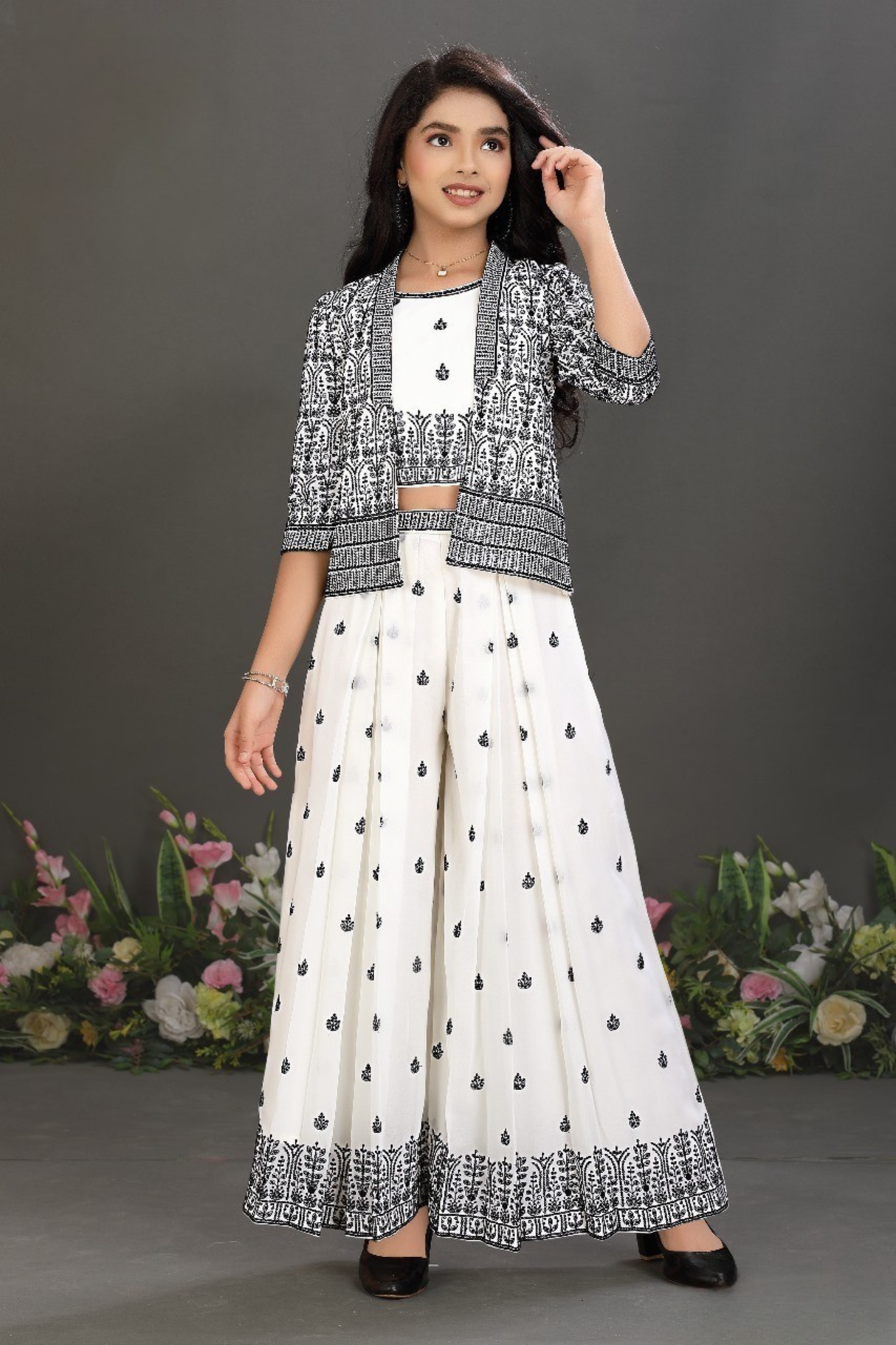 Young girl wearing an elegant cream sharara suit with gold embroidery on the tunic and a matching pleated sharara, styled with a large, colorful floral print shawl.