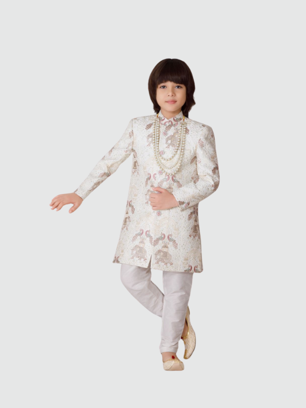 Young boy posing in a full-sleeve cream/off-white sherwani with an intricate gold woven (jacquard) peacock and floral pattern, paired with light cream trousers and a long white pearl necklace