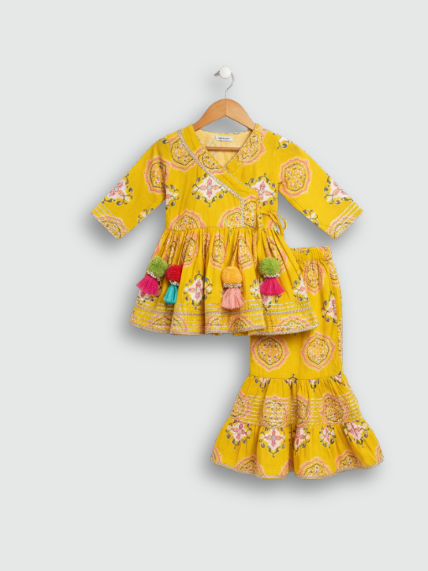 Full set laid flat: a mustard yellow Angrakha-style kurta with pink/white geometric print and multi-colored pompoms, and matching tiered sharara pants.