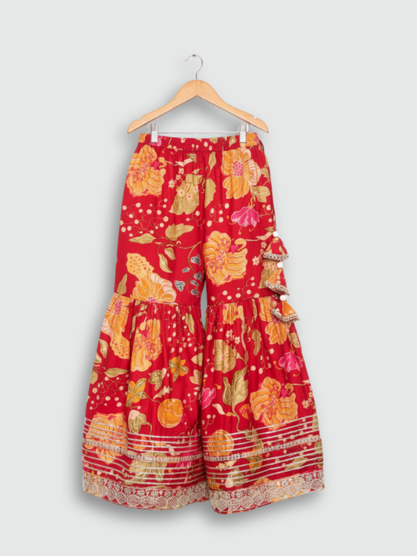 Full set laid flat: a vibrant red, gold-printed kurta with gold embroidered neckline, and matching tiered sharara pants with gold lace detailing.