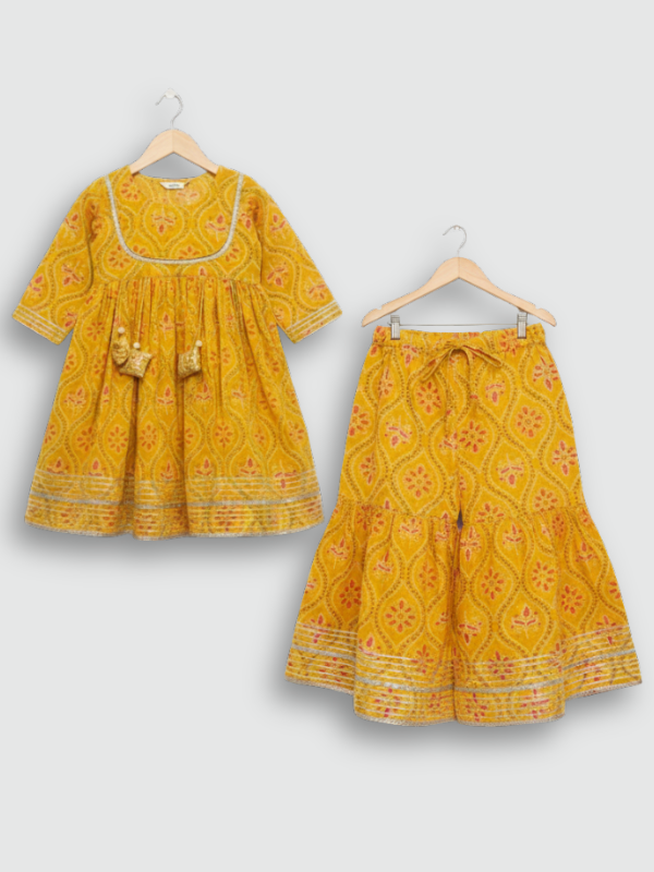 Full set laid flat: a mustard yellow Angrakha-style kurta with pink/white geometric print and multi-colored pompoms, and matching tiered sharara pants.