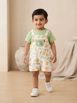 Infant boy wearing a green t-shirt and light-colored printed dungaree shorts with shoulder straps and a front chest pocket.