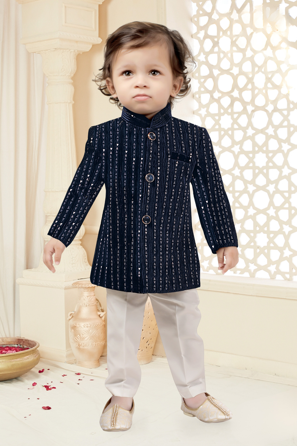  Toddler boy wearing a navy blue velvet short jacket (Jodhpuri style) with vertical silver sequin embroidery, paired with white trousers and cream embroidered ethnic shoes.I 