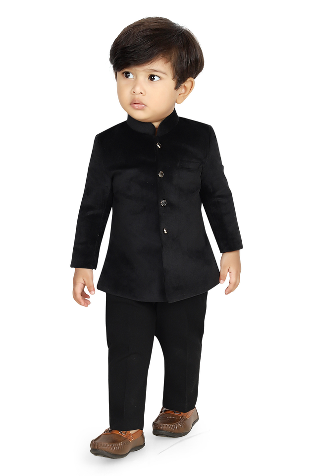  Toddler boy wearing a monochromatic black velvet short jacket (Jodhpuri style) with a mandarin collar, paired with matching black trousers and brown ethnic slip-on shoes.