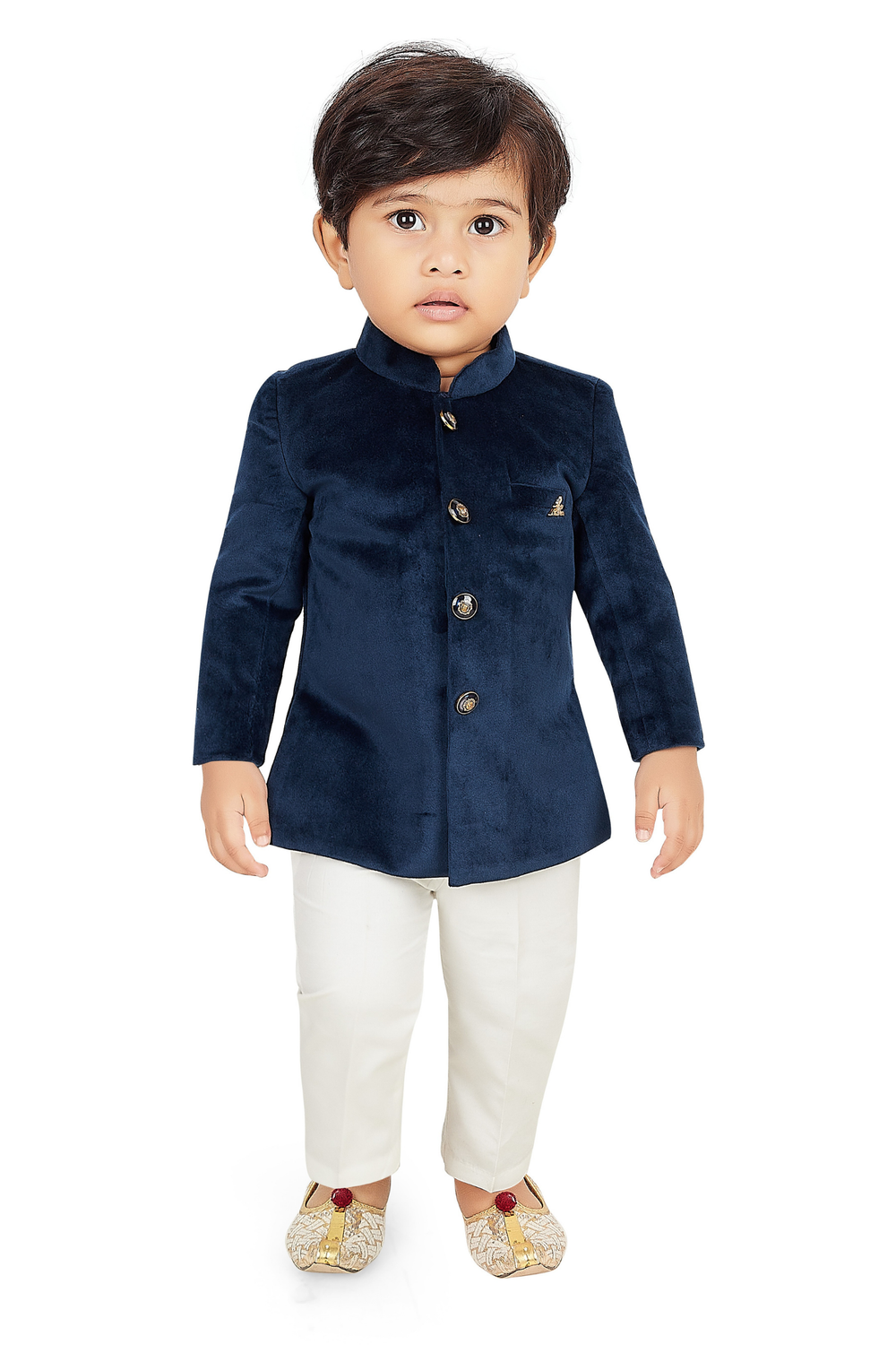  Toddler boy wearing a navy blue velvet short jacket (Jodhpuri style) with gold buttons, paired with cream/off-white trousers and light gold embroidered ethnic shoes.