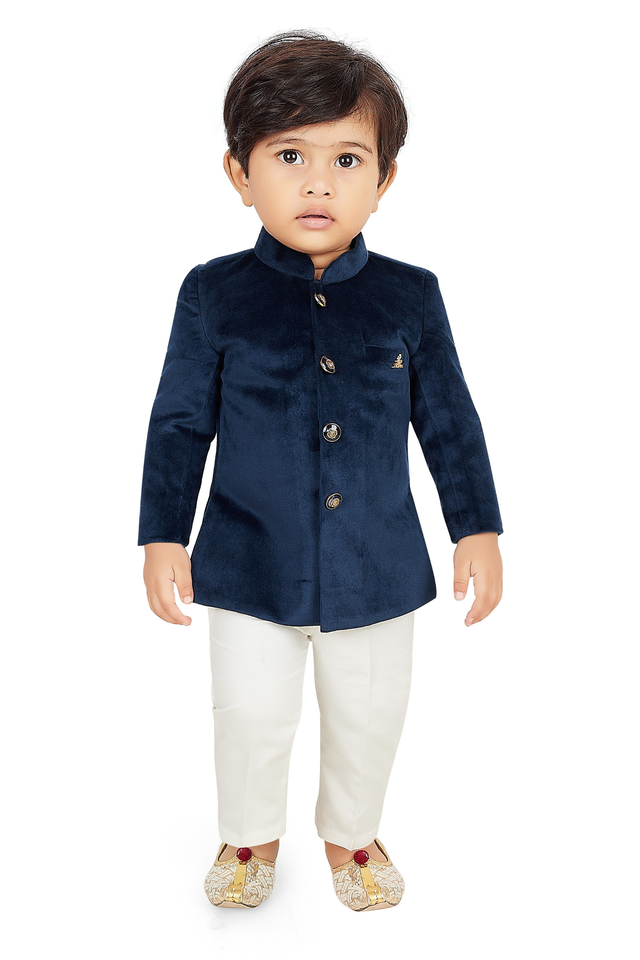  Toddler boy wearing a navy blue velvet short jacket (Jodhpuri style) with gold buttons, paired with cream/off-white trousers and light gold embroidered ethnic shoes.