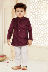 Toddler boy wearing a deep maroon/burgundy velvet short tailored jacket with silver horse embroidery on the chest, paired with cream trousers and gold ethnic shoes.