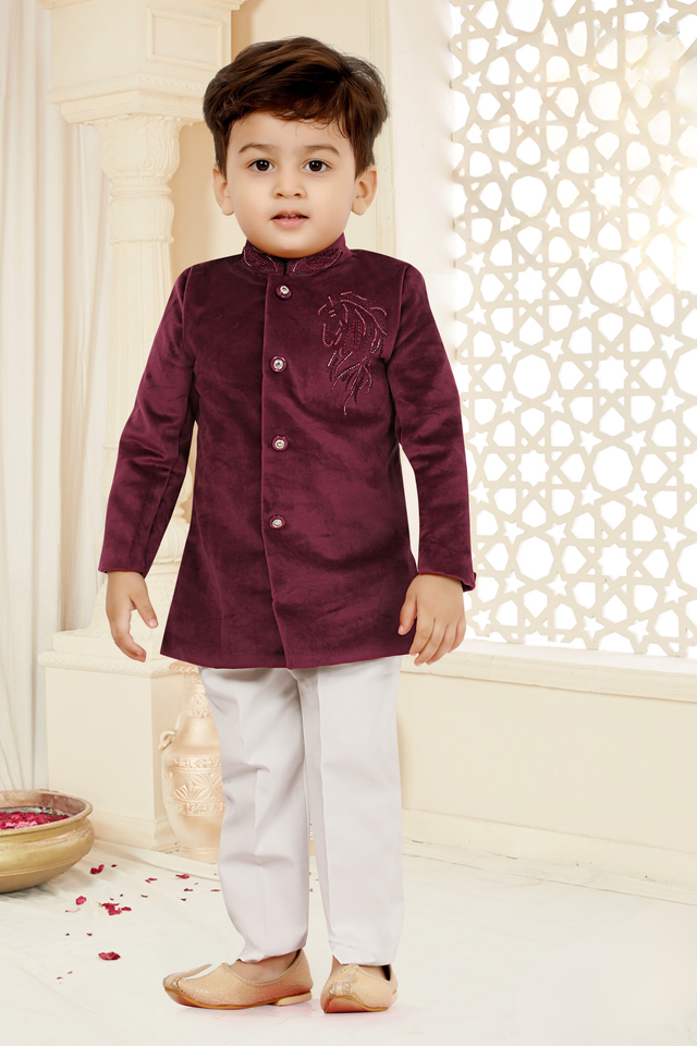 Toddler boy wearing a deep maroon/burgundy velvet short tailored jacket with silver horse embroidery on the chest, paired with cream trousers and gold ethnic shoes.