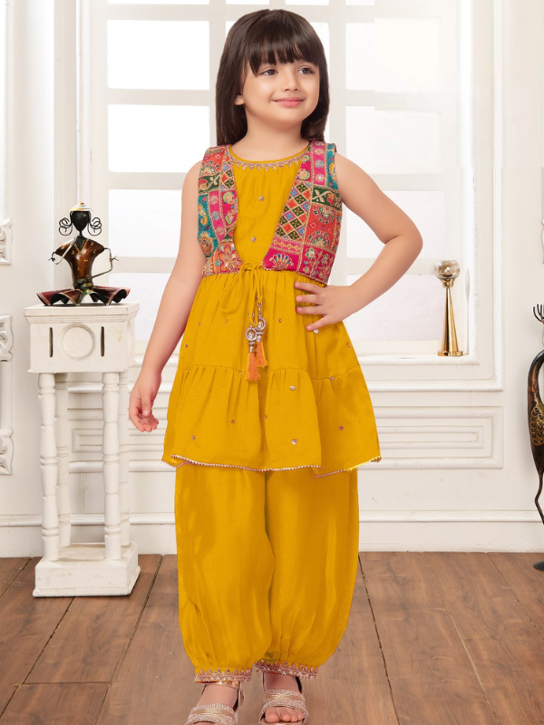 Young girl modeling a bright yellow ethnic wear set with a short, tiered Kurti, balloon-style pants, and a colorful, embroidered sleeveless jacket.
