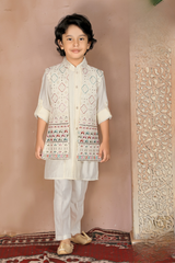 Young boy wearing a fawn-colored kurta pajama set, layered with a sleeveless Nehru jacket featuring a repeated pattern of multi-colored elephants, and light ethnic slip-on shoes.
