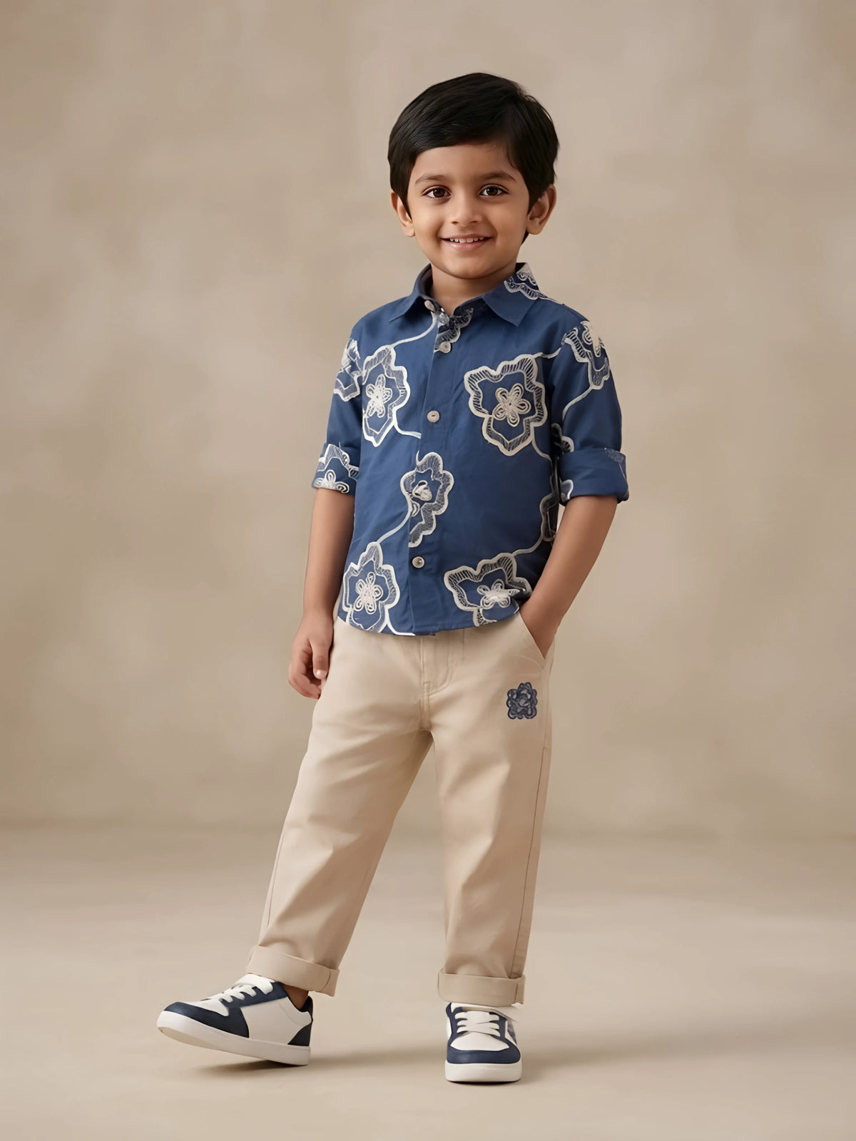 A young boy wearing a navy blue floral printed shirt with rolled-up full sleeves paired with beige trousers and sneakers