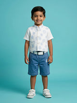 A young boy smiling while wearing a stylish white and blue tropical leaf print waistcoat over a white short-sleeve shirt, paired with washed blue denim shorts, a white belt, and white sneakers against a turquoise background.