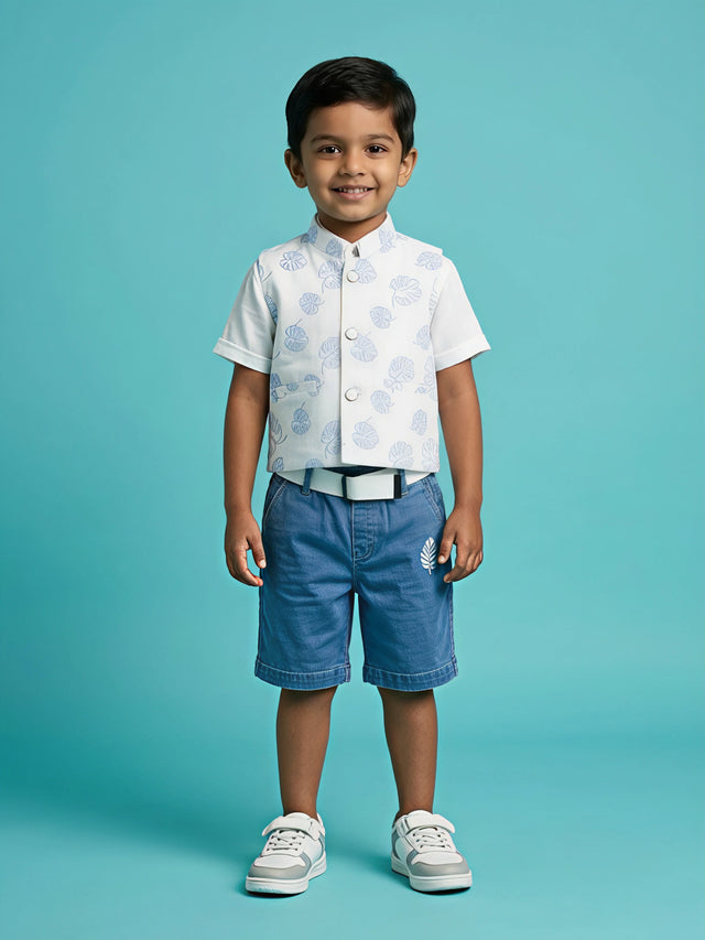 A young boy smiling while wearing a stylish white and blue tropical leaf print waistcoat over a white short-sleeve shirt, paired with washed blue denim shorts, a white belt, and white sneakers against a turquoise background.