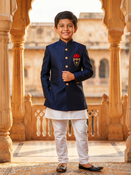 A young boy wearing a premium Navy Blue Jodhpuri Sherwani with gold buttons, a red pocket square, and white trousers in a palace setting.