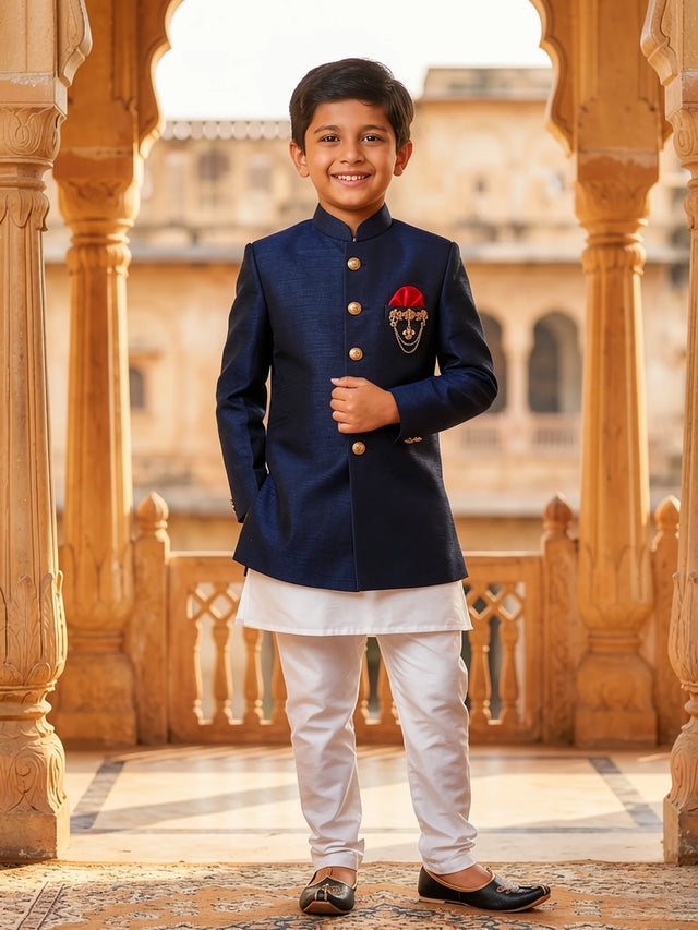 A young boy wearing a premium Navy Blue Jodhpuri Sherwani with gold buttons, a red pocket square, and white trousers in a palace setting.