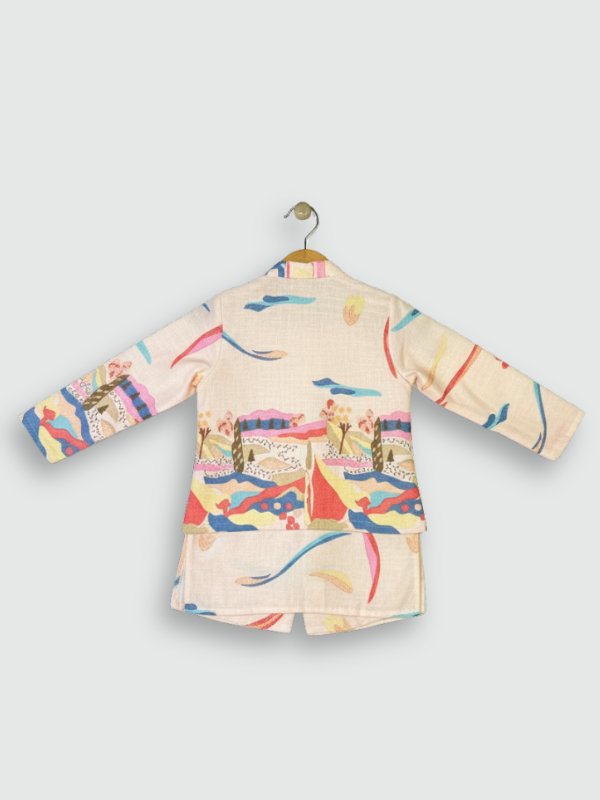 A young boy standing and smiling, wearing a cream long-sleeved kurta and pajama set with a sleeveless, brightly colored abstract-printed Nehru jacket.