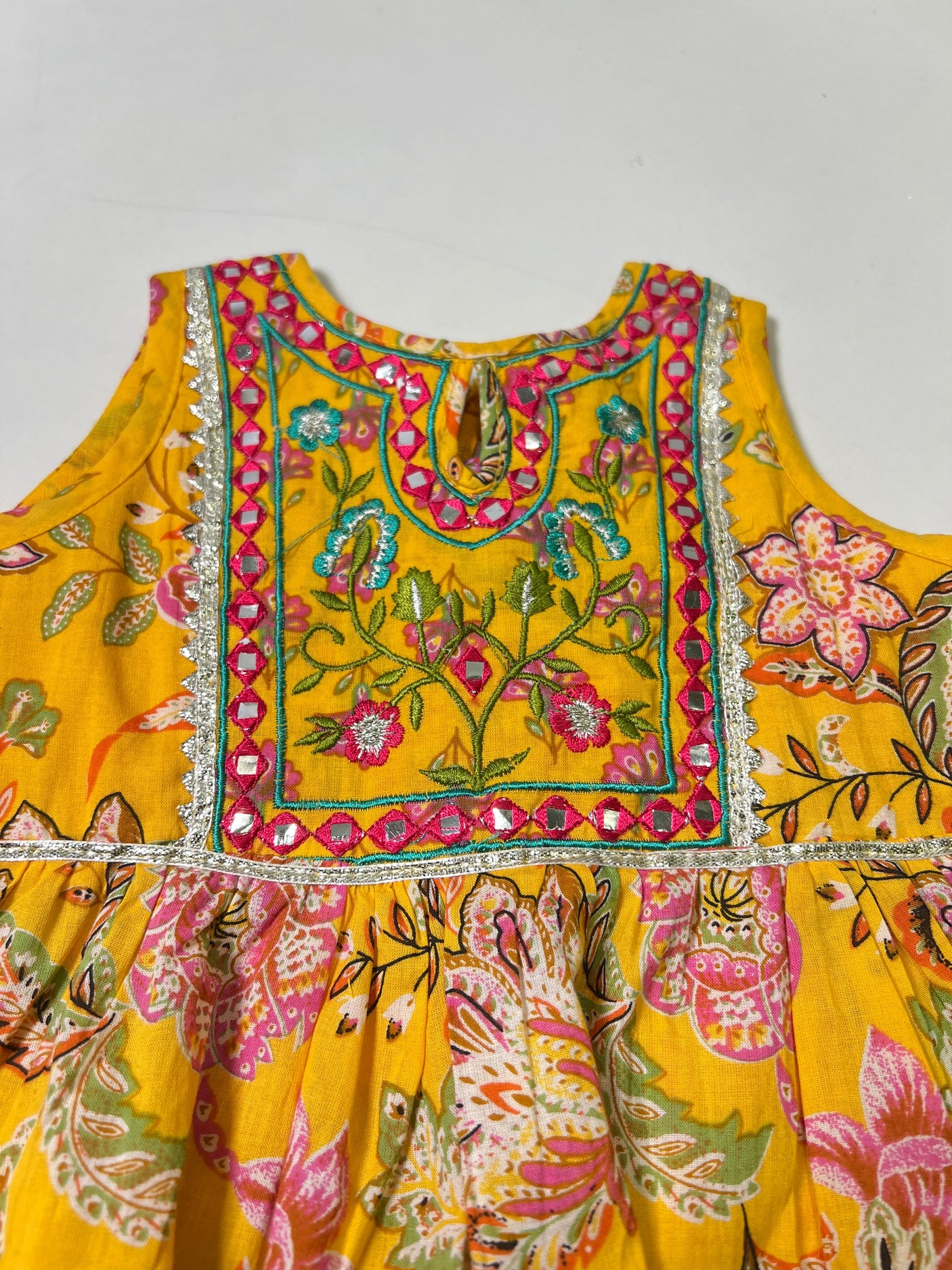 Front view of a girls' sleeveless yellow peplum top with all-over pink/green floral print and a heavily embroidered neckline and yoke, hanging on a wooden hanger.