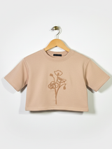 Front view of a girls' nude/tan short-sleeve cropped t-shirt with a thin, contrasting bronze floral outline embroidery on the chest, hanging on a wooden hanger.