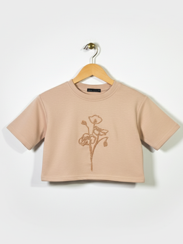 Front view of a girls' nude/tan short-sleeve cropped t-shirt with a thin, contrasting bronze floral outline embroidery on the chest, hanging on a wooden hanger.