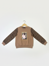 Front view of a girls' brown sweatshirt with long leopard print sleeves and a framed graphic of a girl on the chest, hanging on a wooden hanger.