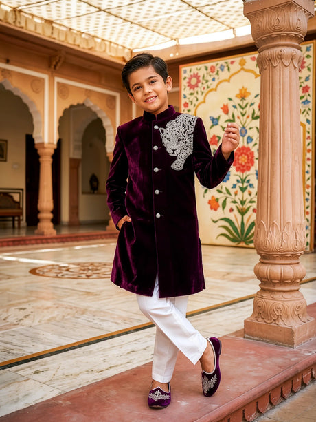 Young boy wearing a royal purple velvet sherwani with silver panther embroidery and white trousers in a traditional palace setting.