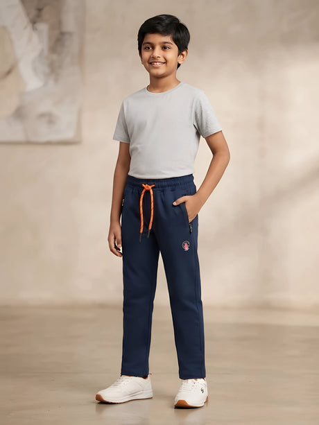 Young boy wearing navy blue slim fit trackpants with orange drawstrings, side zipper pockets, grey t-shirt, and white sneakers.