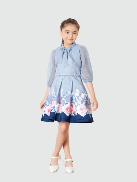 Young girl smiling in a dusty blue party dress with a sheer puff-sleeve shrug, a silver belt, and a flared skirt with a large pink and white rose floral border print