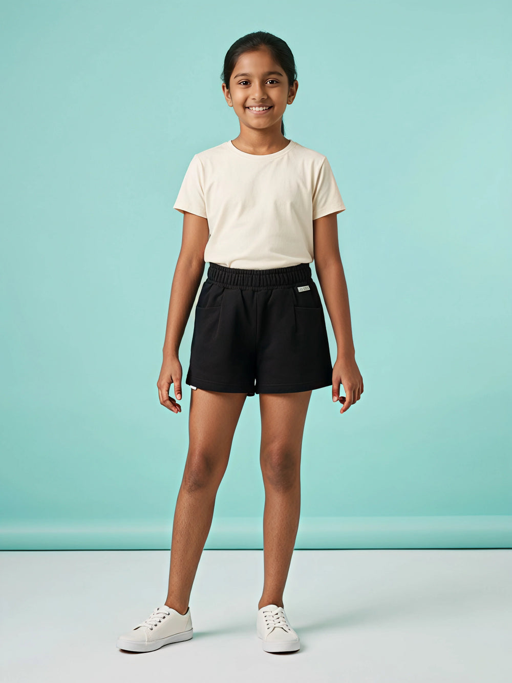 A young girl wearing comfortable solid black shorts with an elastic smocked waistband and front pockets, paired with a cream t-shirt against a light blue background.