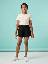 A young girl wearing comfortable solid black shorts with an elastic smocked waistband and front pockets, paired with a cream t-shirt against a light blue background.