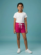 A young girl standing against a blue background wearing bright Rani pink casual shorts featuring a white drawstring and side pockets, paired with a white t-shirt and white sneakers.