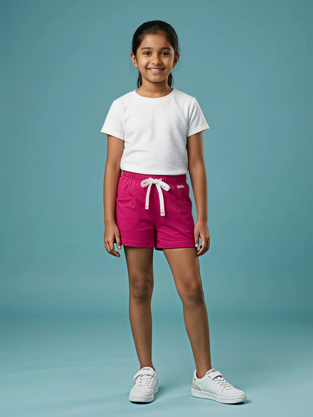 A young girl standing against a blue background wearing bright Rani pink casual shorts featuring a white drawstring and side pockets, paired with a white t-shirt and white sneakers.