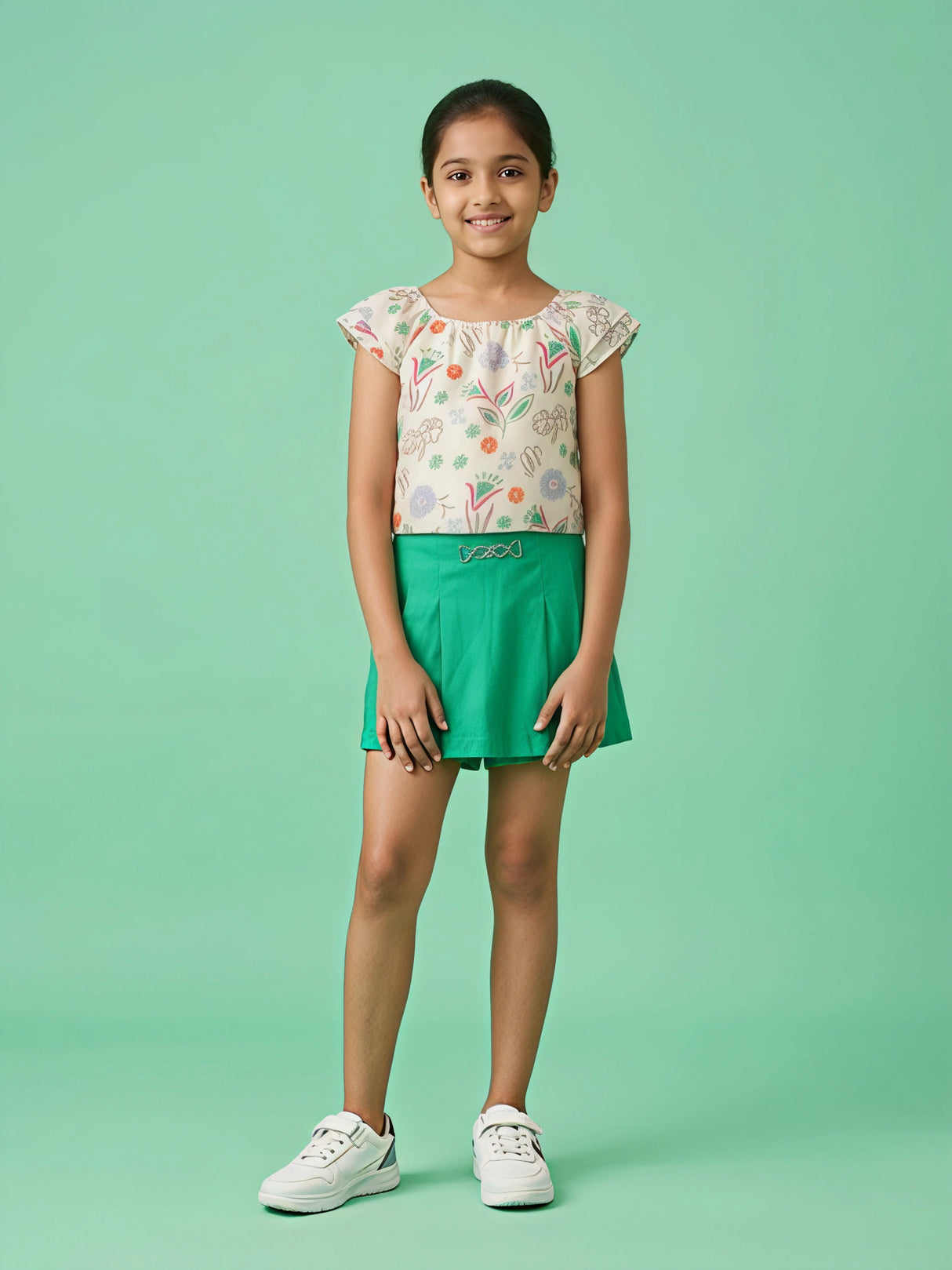 A young girl posing in a two-piece outfit featuring a cream-colored short-sleeve top with colorful floral doodles and a vibrant green short skirt with a silver waist detail.